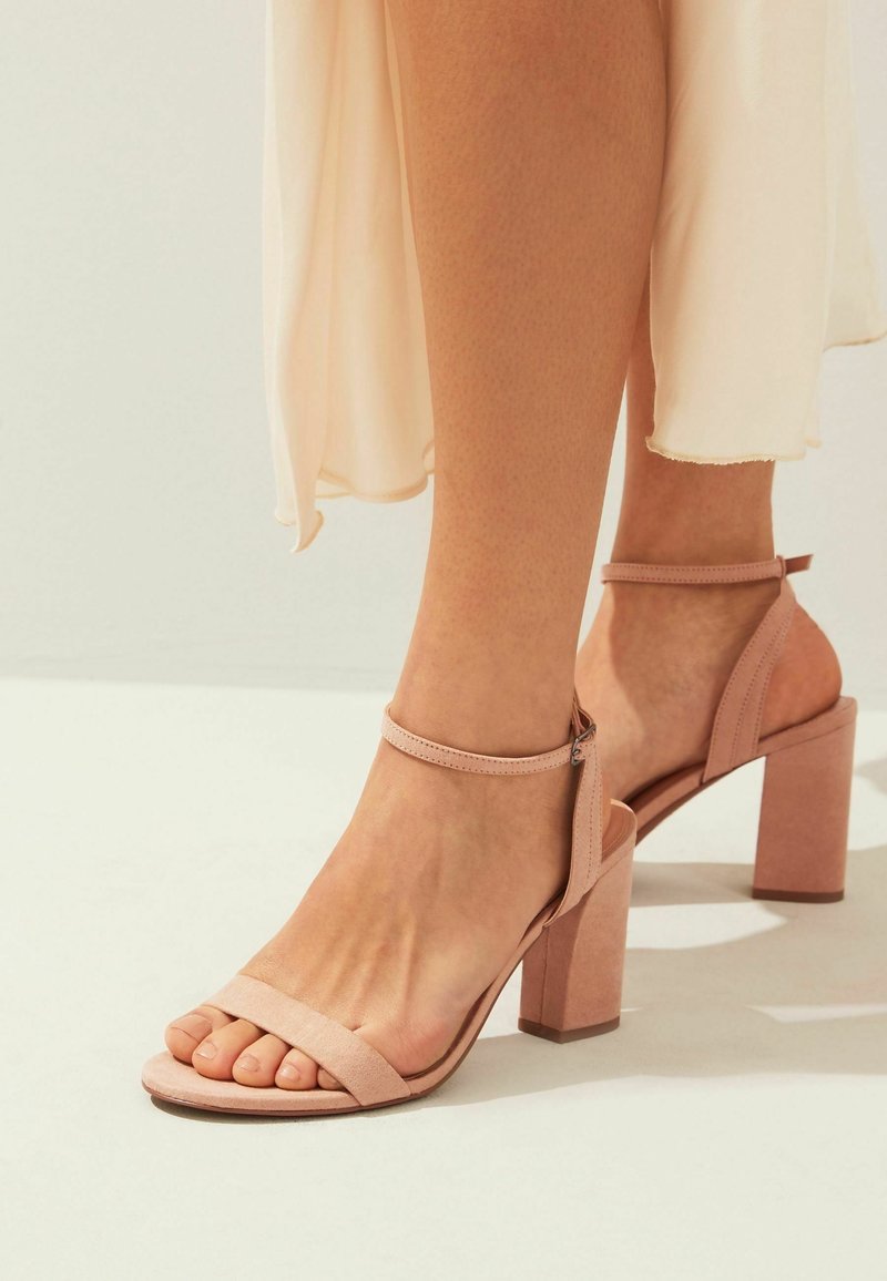 Pieds portant des sandales à talons blocs couleur nude avec des brides autour de la cheville, associés à une jupe légère et fluide.
