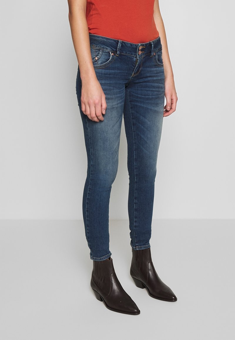 Woman wearing faded blue skinny jeans, dark brown ankle boots, and a sleeveless rust-orange top, standing against a plain background.