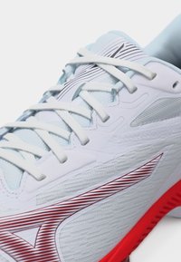 Λευκό αθλητικό παπούτσι με λευκά κορδόνια, επάνω μέρος από υφασμάτινο υλικό με υφή και κόκκινη σόλα που διαθέτει λογότυπο με διαγώνιες ρίγες.