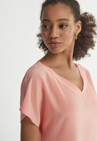 Blusa rosa chiaro con scollo a v realizzata in tessuto morbido, con maniche corte. Presenta un taglio comodo e una drappeggiatura delicata. Nessun motivo o dettaglio visibile.
