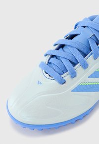 Botas de fútbol azul claro con material sintético texturizado, cordones planos y un vibrante acento en zigzag verde en el costado, con una suela exterior de goma resistente.