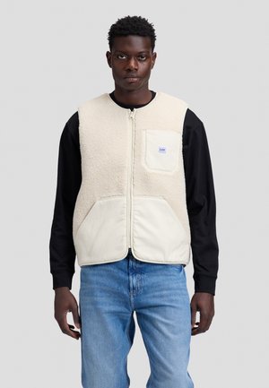 Ung mand iført en cremefarvet fleecevest med frontlynlås over en sort langærmet skjorte og blå jeans, vendt mod fronten mod en ensfarvet baggrund.