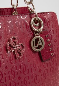 Sac à main rose brillant avec motif logo en relief. Comprend des éléments en métal, y compris une bandoulière chaîne et un porte-clés avec les étiquettes "LA" et "GUESS".