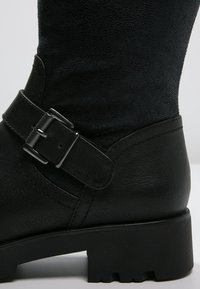 Bota de cuero negro con un acabado texturizado, de altura hasta el tobillo, con una correa con hebilla y una suela gruesa y estriada para mayor tracción.