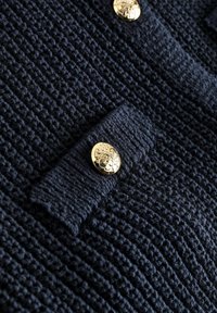 Mornarska teksturirana cardigan jakna s pozlaćenim, uzorkovanim gumbima i prepoznatljivim pletenim dizajnom s preklopnim ovratnikom i bočnim džepovima.