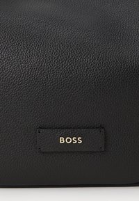 Zwarte leren tas met een gestructureerd oppervlak, met een goudkleurig merklabel "BOSS" op een rechthoekig accentpatch.