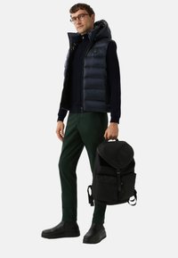 Gepolsterte marineblaue Weste, schwarzes Langarmoberteil, dunkelgrüne Hose, schwarze Tasche mit Klappe und schwarze Schuhe. Lässiges Outfit mit modernem Design.