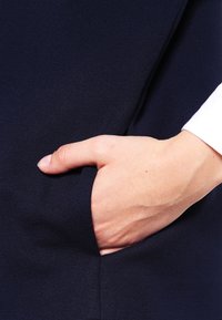 Ruka s viditelným palcem zasunutá do kapsy tmavě námořnického oděvu, na sobě má bílou košili.