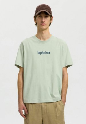 Mladý muž oblečený v svetlozelenom tričku s nápisom „lapiscine“ a béžových nohaviciach, hnedá čiapka, stojí s jednou rukou v vrecku.