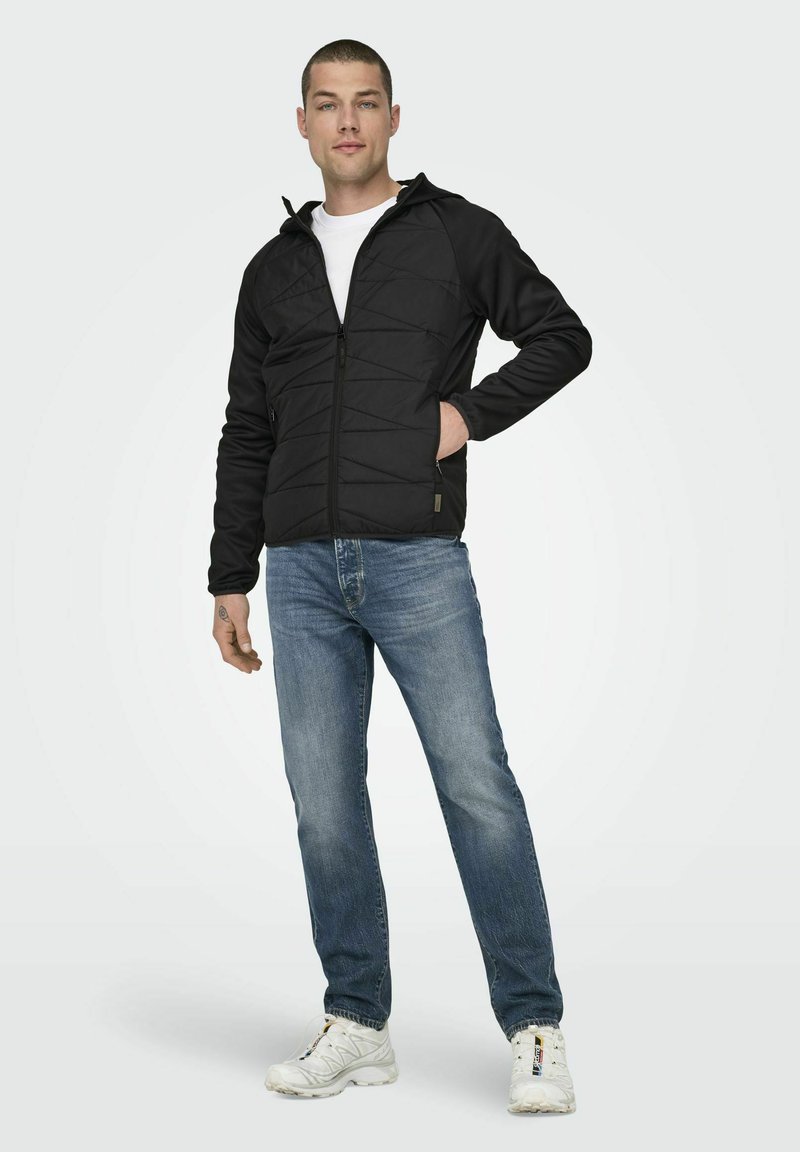 Veste noire matelassée zippée avec capuche, associée à un jean bleu clair. La veste possède des poches latérales et une finition texturée.