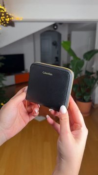 Zwarte leren portemonnee met gouden ritssluiting en logo, rechthoekige vorm, licht gestructureerd oppervlak, in handen gehouden tegen een vage achtergrond.