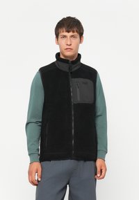 Zwarte fleece vest met ritssluiting aan de voorkant, opstaande kraag en een borstzak. Kenmerkt zich door een gestructureerd materiaal en wordt gedragen over een teal langslavenshirt.