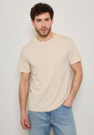 Homme aux cheveux courts et bouclés portant un t-shirt beige à manches courtes et un jean bleu clair, debout devant un fond clair uni.