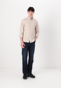 Camisa de botones a rayas beige con mangas largas, jeans azul marino y zapatillas negras. La camisa presenta un cuello clásico y un detalle de bolsillo en el frente.