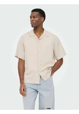 Homme portant une chemise beige à manches courtes boutonnée et un jean déchiré bleu clair, debout avec une main dans la poche, regardant sur le côté.