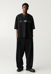 T-shirt nera oversize con stampe grafiche, abbinata a pantaloni larghi neri e scarpe chunky nere, su uno sfondo neutro.