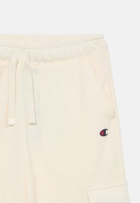 Εκρού φόρμα με ελαστική ζώνη μέσης, κορδόνι, πλαϊνή τσέπη και μικρό λογότυπο Champion στο πάνω μέρος του μηρού.