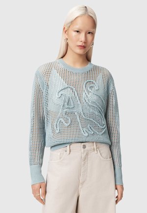 Frau mit langen blonden Haaren, die einen transparenten hellblauen Strickpullover über einem passenden Bralette und hoch taillierte beigen Hosen trägt, stehend vor grauem Hintergrund.