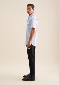 Camicia azzurro chiaro a maniche corte con due tasche sul petto, abbinata a pantaloni scuri e scarpe nere. Design semplice e texture liscia.