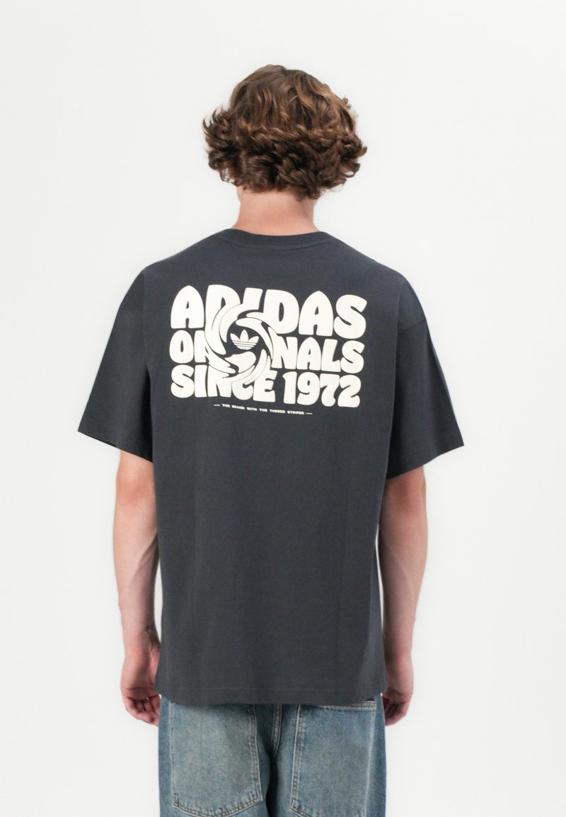 Siva pamučna majica s labavim krojem, s velikim bijelim grafičkim printom "Adidas Originals" na leđima i natpisom "Od 1972." Kratak rukav, okrugli izrez.
