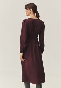 Rochie lungă burgundă cu mâneci lungi, talie elastică, siluetă dreaptă și țesătură fină. Spatele are un guler rotund și mâneci bufante.
