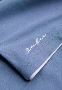 Tissu bleu texturé plié pour montrer une manche avec un texte en cursive brodé en blanc et une bordure blanche le long du bord.