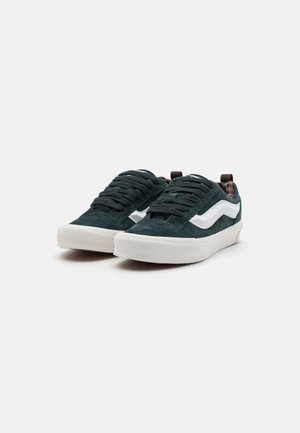 Green suede sneakers with white side stripes, flat laces, padded collar, and a cream rubber sole. Textured material and contrast accents.