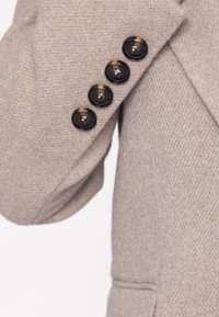 Ärmen på en beige ullkavaj med strukturerat tyg, fyra svarta knappar och diskret sömnadsdetalj.