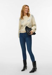 Strikket sweater i creme med et hundemotiv, parret med blå skinny jeans og sorte ankelstøvler. Bærer en sort clutch.