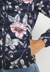 Kukkakuvioinen puhvijakku, jossa on tummansininen kangas, valkoisia ja vaaleanpunaisia kukkakuoseja, vetoketjukiinnitys ja elastiset hihansuut; suunniteltu lämpöä ja tyyliä varten.