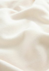 Kermavärinen, pehmeä kangas, jossa on sileä, hieman teksturoitu pinta, esittäen lempeitä poimuja ja hienovaraisia värivaihteluita.