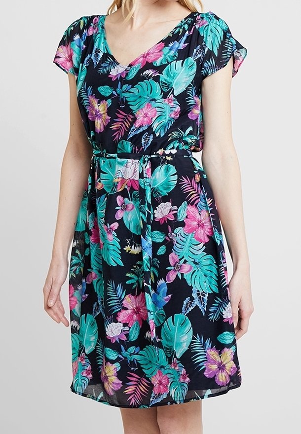 Femme portant une robe noire mi-longue avec imprimé floral tropical coloré et ceinture nouée à la taille.