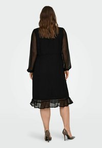Czarna sukienka z długimi transparentnymi rękawami, dopasowaną talią i falbaniastym dołem. Materiał wydaje się lekki o gładkiej teksturze. Nosi ją w połączeniu z brązowymi szpilkami.