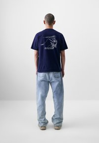 Tricou din bumbac bleu marin, cu un imprimeu grafic pe spate care reprezintă o persoană pescuind. Asortat cu o pereche de blugi lejeri de culoare albastru deschis.