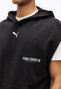 Man draagt een zwarte mouwloze Puma-hoodie met een wit Puma-logo en de tekst "Puma Crew '48" op de borst, over een wit T-shirt.