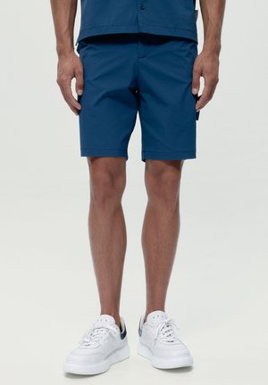 Hombre con pantalones cortos azul marino y zapatillas blancas de pie contra un fondo liso y claro, mostrando las piernas y las manos a los lados.