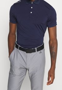 Marinblå pikétröja med tre knappar, korta ärmar och "RLX"-logotyp på axeln. Kombinerad med ljusgrå byxor och ett svart bälte med rektangulär spänne.