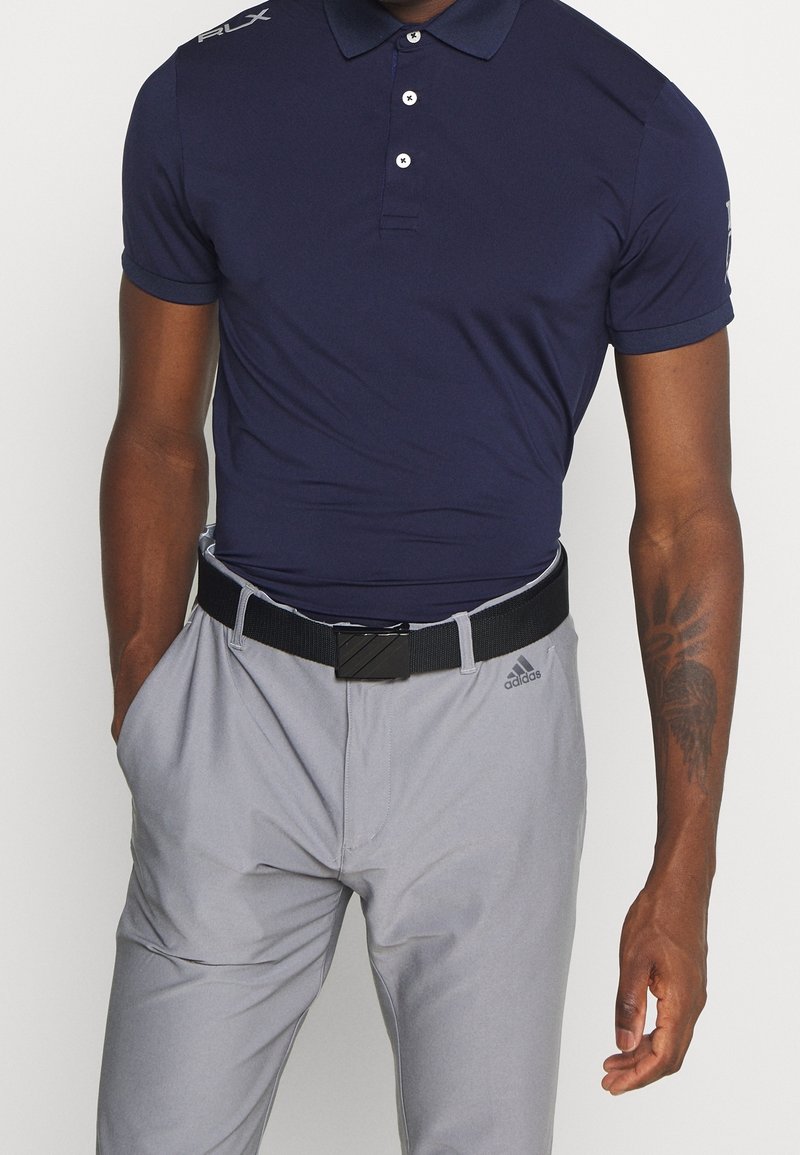 Marinblå pikétröja med tre knappar, korta ärmar och "RLX"-logotyp på axeln. Kombinerad med ljusgrå byxor och ett svart bälte med rektangulär spänne.