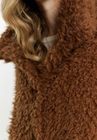 Brun, tekstureret falsk pelsjakke med en bred krave; blødt, fluffy materiale og ujævn overfladebehandling; varmt, hyggeligt udseende.