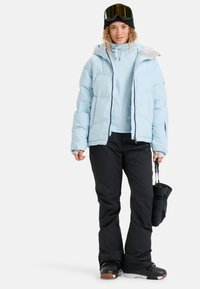 Lichtblauwe pufferjack met capuchon, zwarte skibroek en donkere sneeuwgoggles. Gestructureerde stof en contrasterende ritsaccenten.