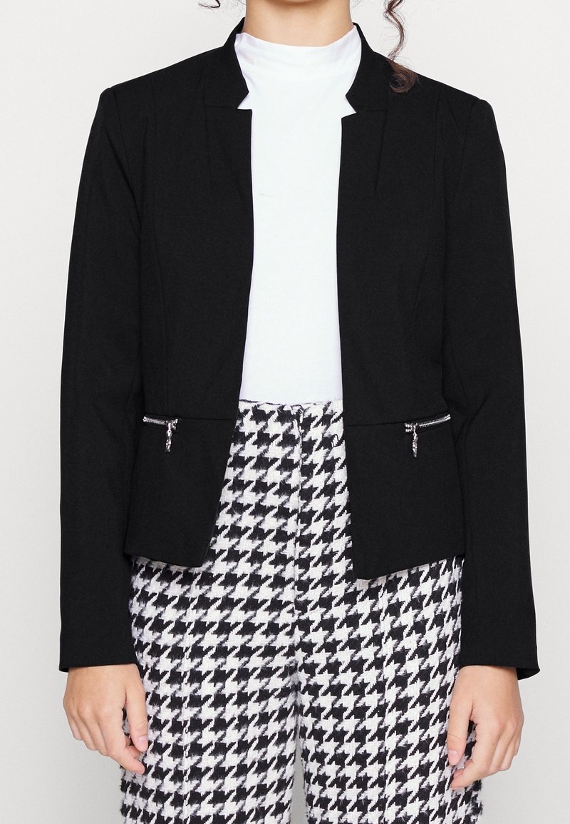 Blazer noir avec une coupe ajustée, doté d'un col cranté et de poches zippées. Porté sur un haut blanc et associé à un pantalon à motif pied-de-poule.