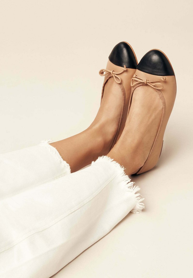 Ballerines en cuir beige avec embout rond noir, détail nœud à l'avant et texture lisse, associées à un pantalon blanc avec ourlet effiloché.