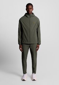 Completo giacca e pantaloni verde oliva, tessuto leggero, chiusura con zip, design con cappuccio, logo giallo sul petto, abbinato a sneakers chiare.