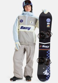 Snowboarder in een grijze en lichtblauwe jas met Roxy-merk, tan sneeuwbroeken, handschoenen en een zwarte snowboard met witte en gekleurde accenten.