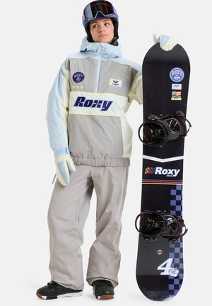 Surfeur des neiges portant une veste grise et bleu clair avec un logo Roxy, un pantalon de neige beige, des gants et un snowboard noir avec des accents blancs et colorés.