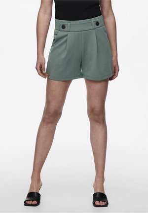 Kvinde iført højtaljede grønne shorts med knapper foran, kombineret med sorte slidesandaler og en sort top, stående med let spredte ben.