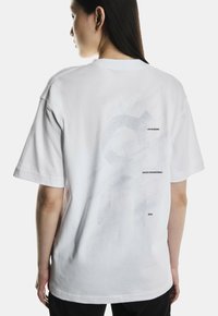 Kvinde med langt mørkt hår iført en løs hvid T-shirt med et svagt prikket mønster og lille sort tekst, der står "ON RUNNING," "SWISS ENGINEERING," og "2010" på ryggen.