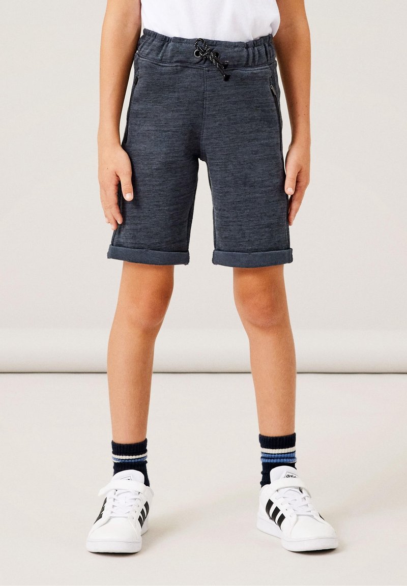 Enfant portant un short gris foncé avec revers, des baskets blanches avec des rayures noires et des chaussettes rayées bleu marine, debout devant un fond uni.