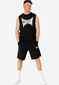 Fekete ujjatlan póló fehér "TAPOUT" logóval, fekete bermuda shortssal, amelyen szintén ugyanaz a logó található, fehér sportcipőkkel és zoknikkal kiegészítve.