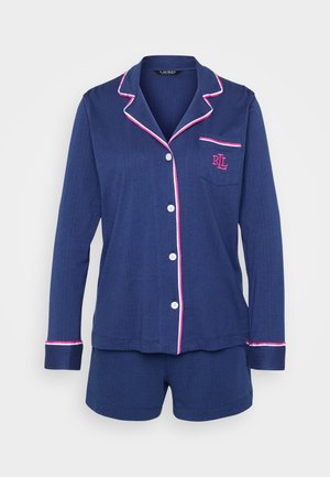 Conjunto de pijama em azul marinho com uma camisa de botões e calções. Apresenta um acabamento em branco e rosa ao longo do colarinho, punhos e bolso com o logótipo.